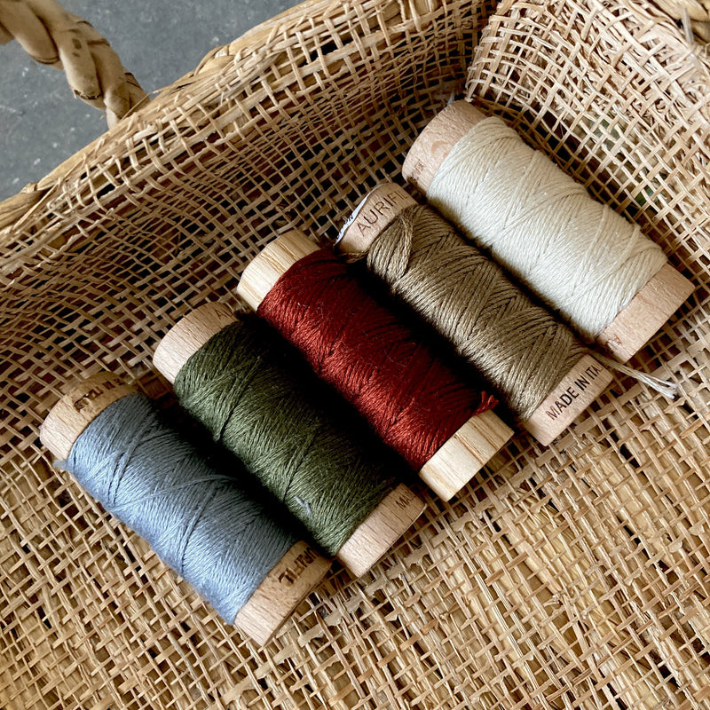 A basket holding 5 colors of embroidery floss on wooden spools - the colors include gray, forest green, maroon, tan, and cream.