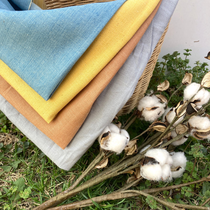 four colors of denim fabric folded and hanging out of a wicker basket next to dried cotton branches and atop green grass